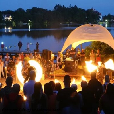 Widoczne osoby tańczące taniec z ogniem, dookoła publiczność. W tle scena z namiotem i jezioro.