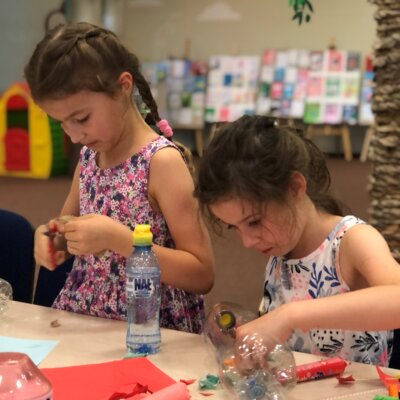 Dwie dziewczynki wykonują statek z plastikowej butelki. Używają nożyczek, plasteliny i skrawków papieru.