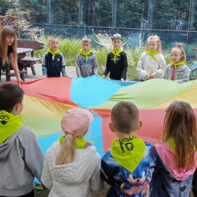 Grupa dzieci wraz z Panią bibliotekarką trzyma kolorową chustę animacyjną. Kliknięcie powoduje powiększenie zdjęcia