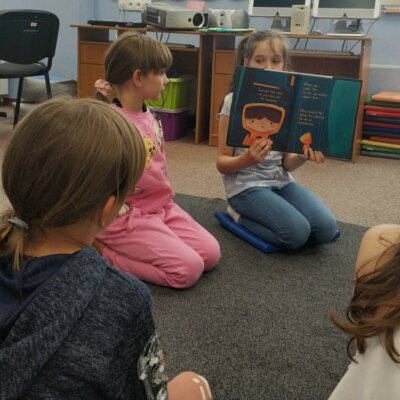 Dzieci siedzą na dywanie, patrzą w stronę dziewczynki, która pokazuje im ilustracje w książce.. kliknięcie powoduje powiększenie zdjęcia