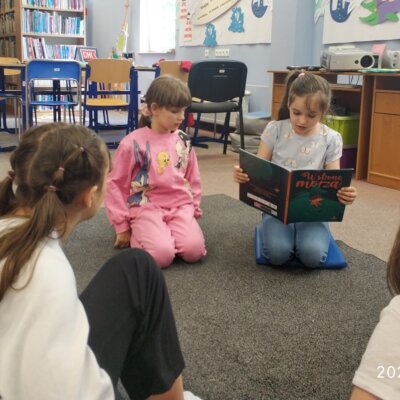 Dzieci siedzą na dywanie i słuchają tekstu czytanego przez dziewczynkę w kitce.. Klikniecie powoduje powiększenie zdjęcia