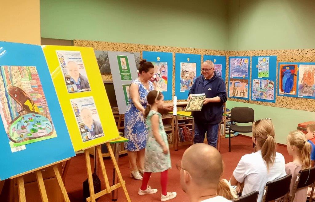 Ośmioletnia Laureatka odbiera nagrodę. Pisarz- Marcin Pałasz podaje dziewczynce książkę. W tle widzimy nagrodzone prace. Po prawej stronie są na sztalugach. Po lewej rysunki wiszą na ścianach. Kliknięcie powoduje powiększenie zdjęcia