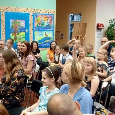 Dzieci i dorośli mają uśmiechnięte twarze. Uczniowie po prawej stronie podnieśli dłonie. Zadają pytania pisarzowi. Za nimi są regały z książkami.. Klikniecie powoduje powiększenie zdjęcia
