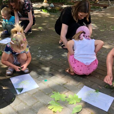 Troje dzieci wraz z rodzicami maluje za pomocą liścia koronę drzew na kartce. Kliknięcie powoduje powiększenie zdjęcia