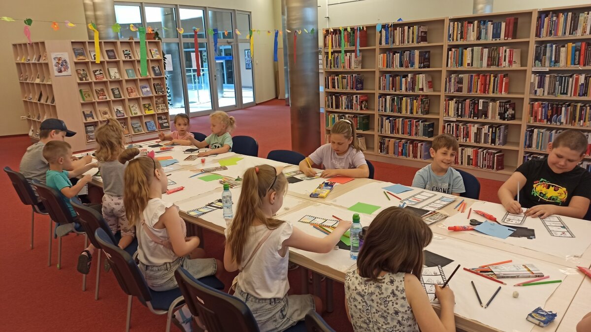 Dzieci siedzą przy stole i malują, w tle regały biblioteczne