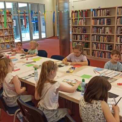 Dzieci siedzą przy stole i malują, w tle regały biblioteczne