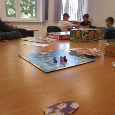 Dzieci grające w gry planszowe przy stołach. Na pierwszym planie widać opakowania od gier: "Boom, Boom śmierdziaki", "Monopoly" i "Dixit" wraz z planszą. Kliknięcie powoduje powiększenie zdjęcia