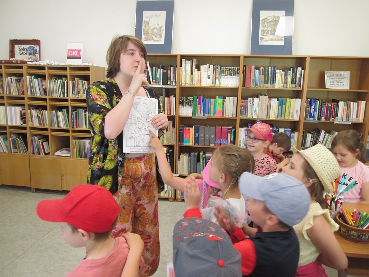 Bibliotekarka z uciszającym placem na buzi, pokazująca grupie dzieci obrazek z basenem "Słowianka" w Gorzowie Wielkopolskim. Kliknięcie powoduje powiększenie zdjęcia