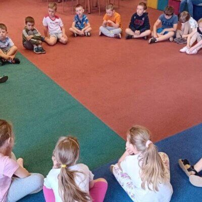 Przedszkolaki siedzą na podłodze w kręgu. Bibliotekarka czyta książkę. Również siedzi.
