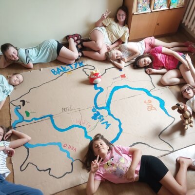 Leżąca na podłodze mapa Polski, przedstawiająca główne rzeki i akweny wodne Polski. Leżące wokół mapy dziewczynki.