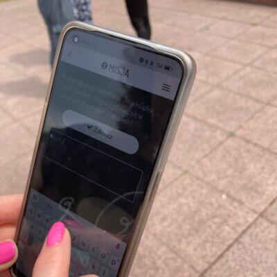 Smartfon w dłoni kobiety. Na ekranie wyświetlony element gry mobilnej. Kobieta wpisuje tekst na klawiaturze smartfonu.