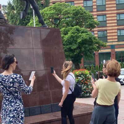 Kobiety ze smartfonami w dłoniach stoją przy pomniku Józefa Piłsudskiego siedzącego na koniu. W tle budynek hotelu oraz zaparkowane samochody.