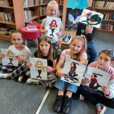 Dzieci siedzące na dywanie prezentują pokolorowane „rosnącymi farbami” ilustracje postaci z warszawskich legend