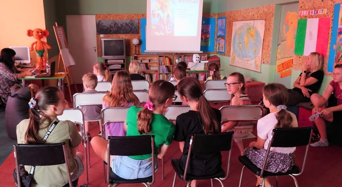 Grupa dzieci ogląda pokaz slajdów o Meksyku, a podróżniczka opowiada. Kliknięcie powoduje powiększenie zdjęcia.