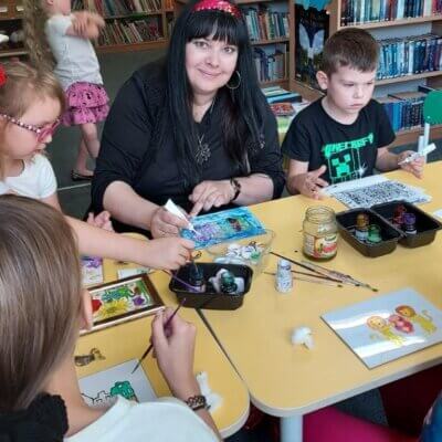 Iwona Mazur, gorzowska artystka plastyczka prowadząca zajecia pomaga dzieciom siedzącym przy stolikach w malowaniu farbami na szklanych taflach