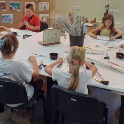 Dziesięcioro dzieci siedzi przy stole. Dzieci trzymają w dłoniach papierowe rurki z gazety, którymi wyklejają szablony tekturowych ramek