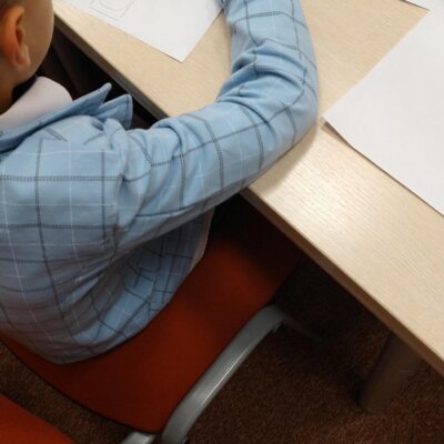 Chłopiec z krótkimi, blond włosami, w niebieskiej koszuli w kratę, siedzi pochylony przy stole, koloruje malowankę niebieską kredką. Kliknięcie powoduje powiększenie zdjęcia.