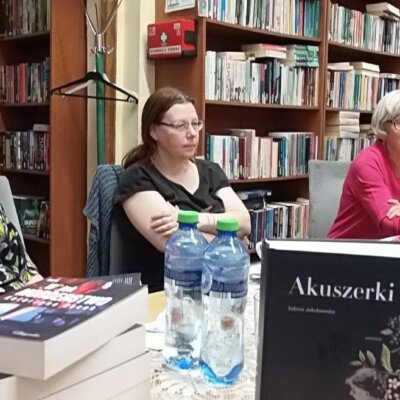 Na pierwszym planie omawiana książka „Akuszerki” oraz stos książek przygotowanych na kolejne spotkania DKK. Przy stole siedzą trzy rozmawiające kobiety. W tle regały wypełnione książkami. Kliknięcie powoduje powiększenie zdjęcia.