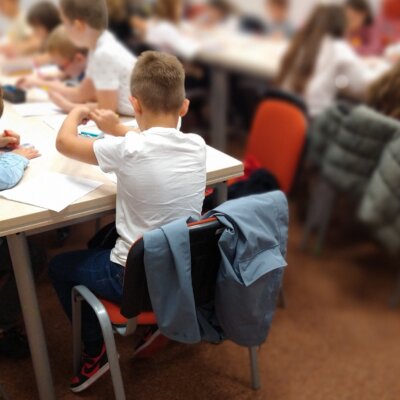 Uczniowie siedzą przy dwóch długich stołach i kolorują malowanki kredkami, zdjęcie jest rozmazane, na pierwszym planie wyostrzone są sylwetki dwóch chłopców siedzących tyłem do obiektywu. Kliknięcie powoduje powiększenie zdjęcia.