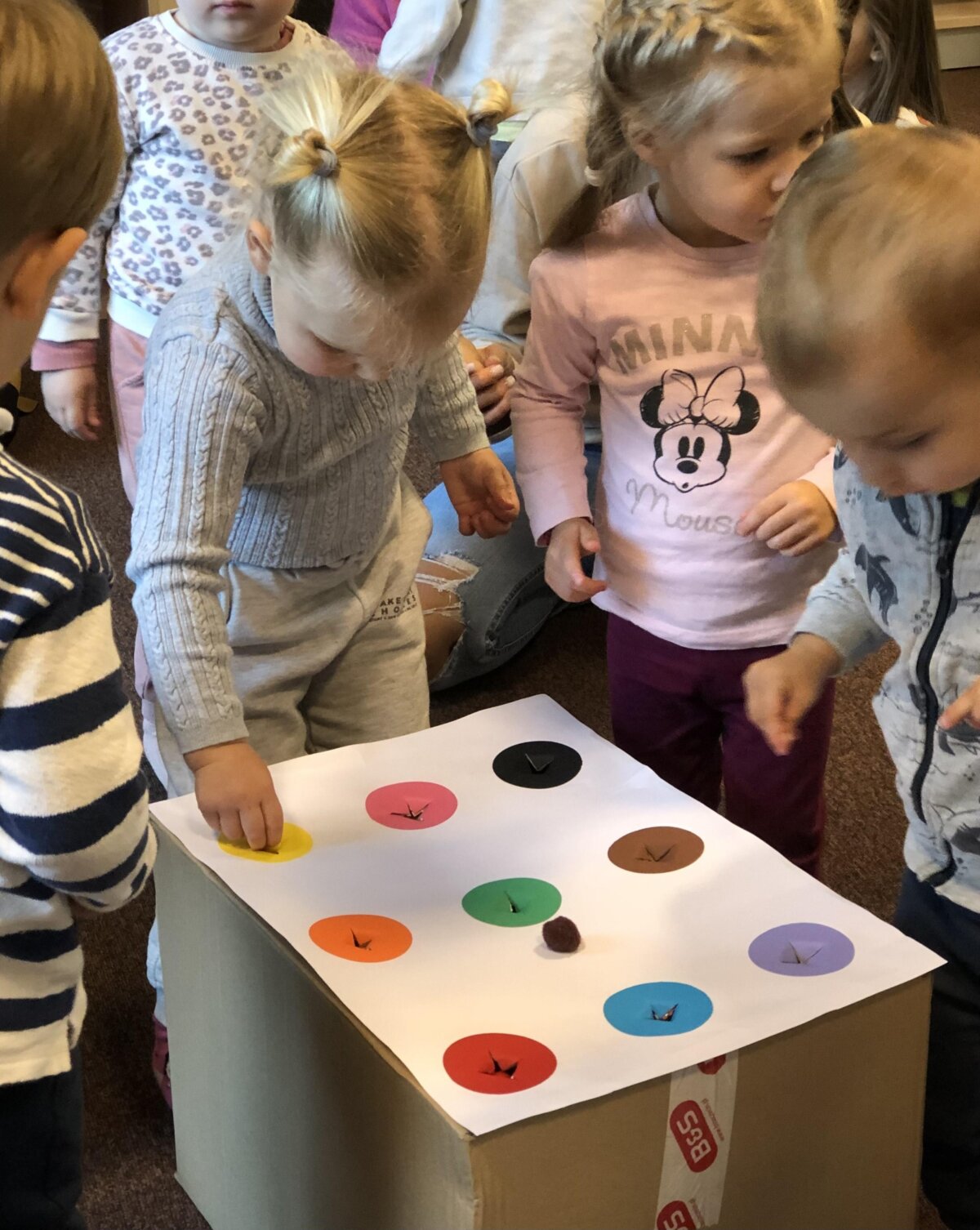 Dzieci wrzucają kolorowe pomponiki do odpowiednich otworów w kartonie.