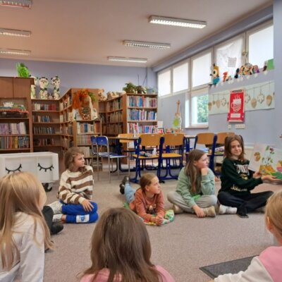 Dzieci siedzą na dywanie, jedna dziewczynka czyta pozostałym dzieciom i pokazuje ilustracje. Kliknięcie w obrazek spowoduje powiększenie.