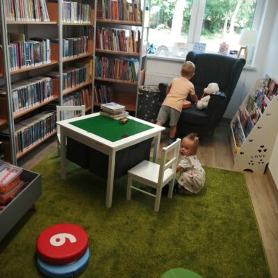 Dziecko siedzi na dywanie, drugie stoi koło fotela. Pod ścianą stoją regały z książkami. Kliknięcie w obrazek spowoduje powiększenie.