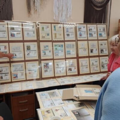 Na zdjęciu widać karty ze znaczkami oprawione w ramy, oparte o ścianę, obok filatelista-starszy mężczyzna stoi bokiem i dwie panie przyglądają się.. Kliknięcie powoduje powiększenie zdjęcia.