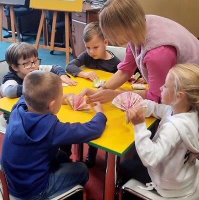 Dzieci przy stole składają serwetki. Nauczycielka pomaga im. Klikniecie powoduje powiększenie zdjęcia.