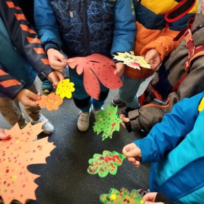 Kilkoro dzieci stojących w kręgu pokazuje swoje prace- liście wycięte z kolorowego papieru i ozdobione farbami w kolorach jesiennych. Kliknięcie powoduje powiększenie zdjęcia.