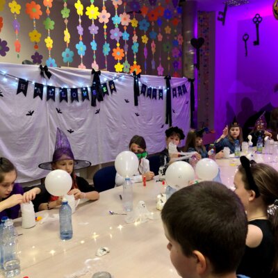 Grupa dzieci siedzących przy stole wykonuje eksperyment przy użyciu balona, sody i octu. Balony w kształcie duchów pompują się wskutek zachodzącej reakcji. Klikniecie powoduje powiększenie zdjęcia.