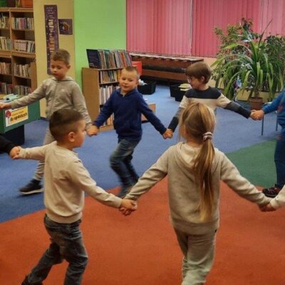 Dzieci i prowadząca trzymają się za ręce. Tańczą wesoło w kółku. Kliknięcie powoduje powiększenie zdjęcia.