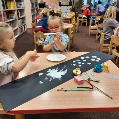 Na pierwszym planie dwie dziewczynki malują białą farbą na czarnej kartce. Jako narzędzia malarskiego używają balona, widelca, jabłka, gąbki i patyka. W tle widać inne dzieci, które wykonują to samo zadanie. Kliknięcie powoduje powiększenie zdjęcia.