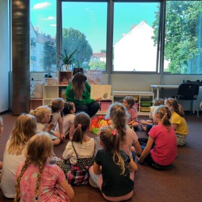 Grupa dzieci siedząca na dywanie słucha książki czytanej przez Panią Bibliotekarkę. Kliknięcie powoduje powiększenie zdjęcia.