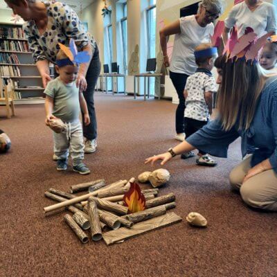 Dzieci przynoszą kamienie i patyczki na ognisko. Pani Bibliotekarka pomaga ułożyć im palenisko.