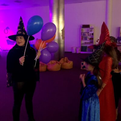 Pani Bibliotekarka w kapeluszu i pelerynie czarownicy trzyma balony, które dzieci przekłuwają. Wewnątrz balonów ukryte są psikusy i cukierki. Klikniecie powoduje powiększenie zdjęcia.