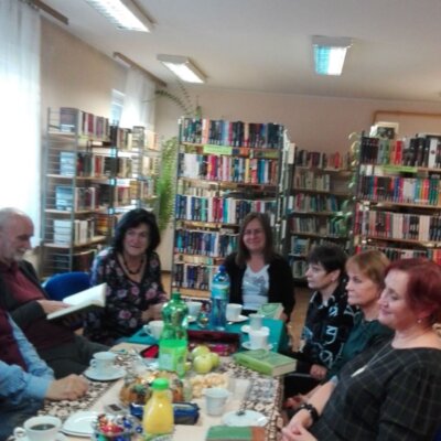 Klubowicze przy stole z książką Elif Shafak „Wyspa zaginionych drzew”. Na stole filiżanki, słodycze, jabłka. Kliknięcie w obrazek spowoduje powiększenie.