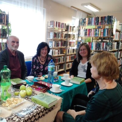 Klubowicze przy stole z książką Elif Shafak „Wyspa zaginionych drzew”. Na stole filiżanki, słodycze, jabłka. Kliknięcie w obrazek spowoduje powiększenie.