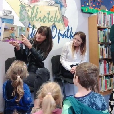 Dwie kobiety, jedna pokazuje ilustracje w książce i dzieci słuchające bajki. Kliknięcie powoduje powiększenie zdjęcia.