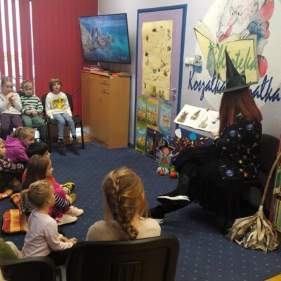 Dzieci siedzą na podłodze, a kobieta w przebraniu czarownicy na krześle czyta bajkę. Kliknięcie powoduje powiększenie zdjęcia.