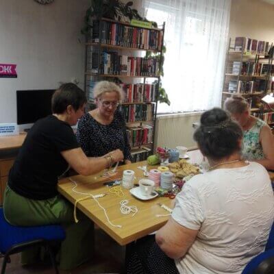 1. Trzy uczestniczki zajęć siedzące przy stole z herbatą, ciastem, sznurkami, nożyczkami, taśmą. Klikniecie powoduje powiększenie zdjęcia.