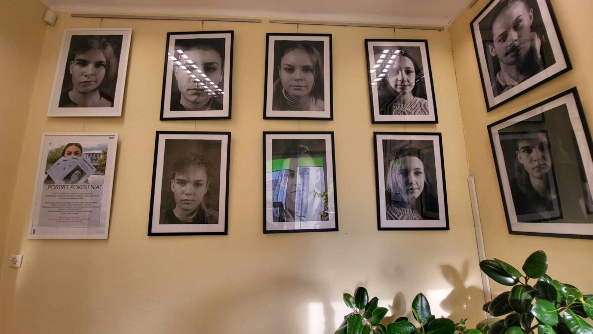 Na żółtej ścianie czarno-białe portrety w czarnych i białych antyramach, plakat prezentujący wystawę w białej antyramie na nim zasłonięta katalogami wystawy twarz dziewczyny, w prawym dolnym rogu zielony fikus. Kliknięcie powoduje powiększenie zdjęcia.