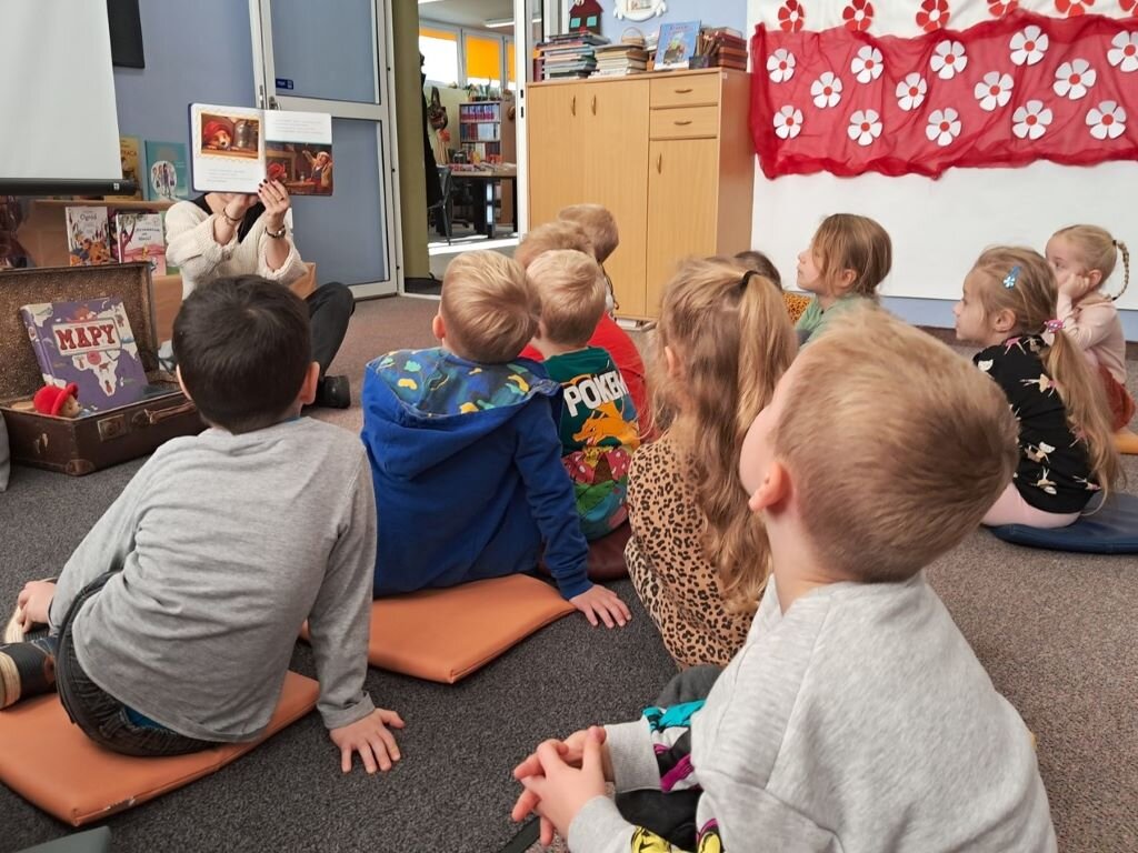 Dzieci siedzą na dywanie, bibliotekarka czyta dzieciom książkę i pokazuje ilustracje.