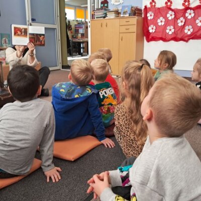 Dzieci siedzą na dywanie, bibliotekarka czyta dzieciom książkę i pokazuje ilustracje.