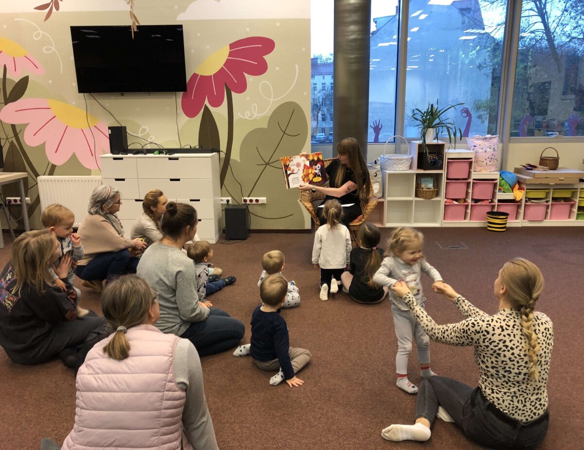 Dzieci wraz z opiekunami słuchają książki czytanej przez Panią bibliotekarkę.