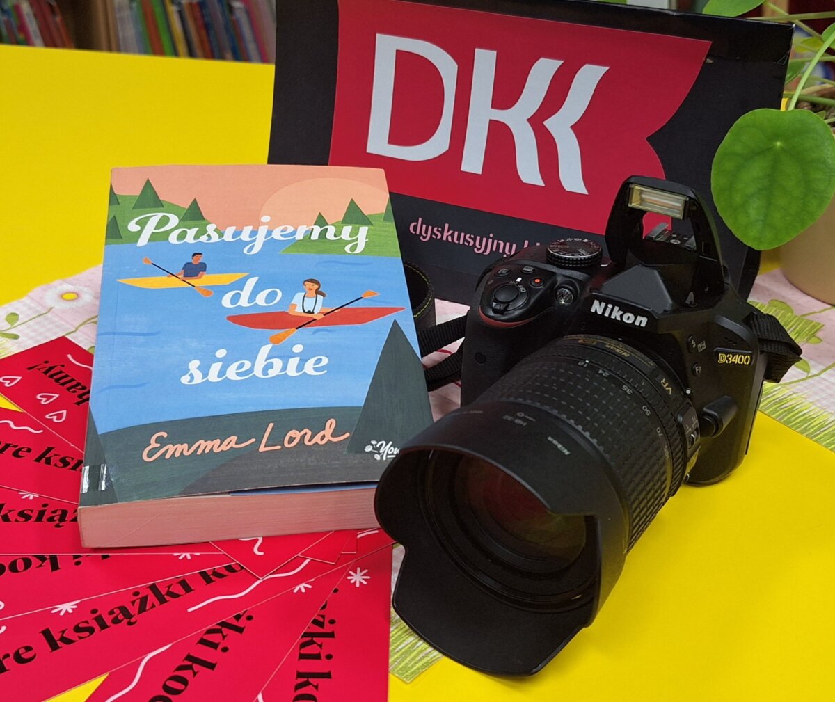 Na stole po prawej stronie leży książka „Pasujemy do siebie” Emmy Lord, a po lewej stronie aparat fotograficzny. Za nimi stoi tabliczka z napisem DKK.