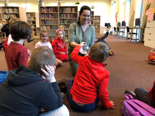 Uśmiechnięte przedszkolaki siedzą na dywanie w okręgu. Pani Bibliotekarka podchodzi do nich z losami, za chwilkę dzieci będą odgrywały wylosowane role.