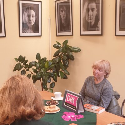 Cztery kobiety siedzące przy stole. Na stole ciemno zielona serweta, emblematy DKK, filiżanki, słodycze. W tle duży zielony kwiat, na jasnej ścianie 5 czarno-białych portretów.