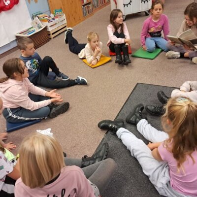 Dzieci siedzą w kole na dywanie, jedna z uczestniczek czyta dzieciom omawianą pozycję. Kliknięcie powoduje powiększenie zdjęcia.