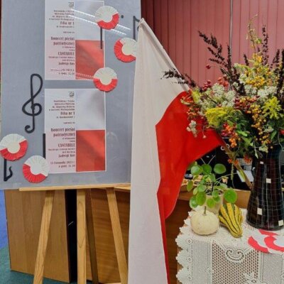 5.Po lewej stronie zdjęcia na sztaludze umieszczony jest plakat zapowiadający koncert. Po środku znajduje się flaga Polski. Z prawej strony na stoliku stoi wazon z jesiennym bukietem, a przed nim leżą ozdobne dynie. Stolik i sztaluga ozdobione są biało-czerwonymi kotylionami.