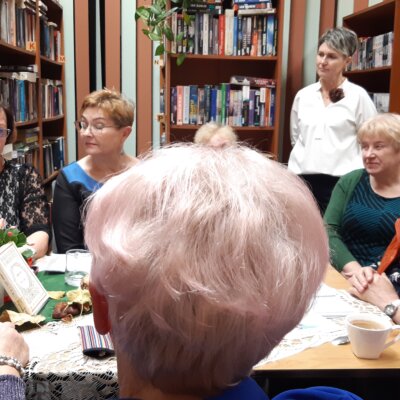 6-Osoby zgromadzone wokół stołu skupiają swój wzrok na prowadzącej spotkanie, która z zaangażowaniem opowiada. Za siedzącymi widać regały z książkami i jeszcze jedną uczestniczkę spotkania stojącą obok nich.